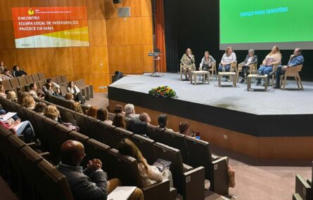 Fotografia de auditório amplo. Na zona do público várias dezenas de pessoas sentadas. No palco, igualmente sentadas (junto de três pequenas mesas) estão seis pessoas.