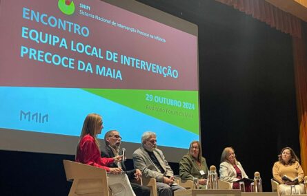 Seis pessoas sentadas, em palco, junto de três pequenas mesas. Uma das pessoas segura um microfone enquanto outra aparenta estar a falar. No fundo do palco, em projeção multimédia, imagem do Encontro da Equipa Local de Intervenção Precoce da Maia.