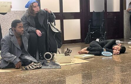Três pessoas com indumentária específica do que aparenta ser uma apresentação teatral. No chão está uma caixa e um par de sapatos (ou sapatilhas).