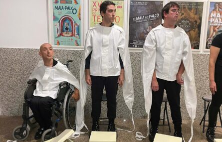 Três pessoas alinhadas. Duas das pessoas estão em pé e uma outra usa cadeira de rodas. Na parede, atrás das pessoas, vários quadros e cartazes afixados.