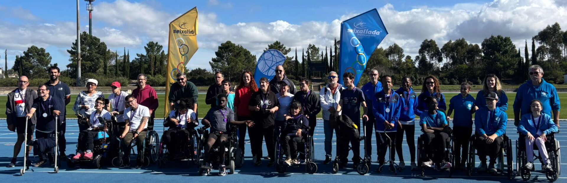 Mais de três dezenas de pessoas alinhadas, lado a lado, em pista exterior do que aparenta ser um complexo de atletismo. Ao fundo imensas árvores. Atrás do grupo estão duas bandeiras (estandartes) com a indicação escrita Seixalíada.