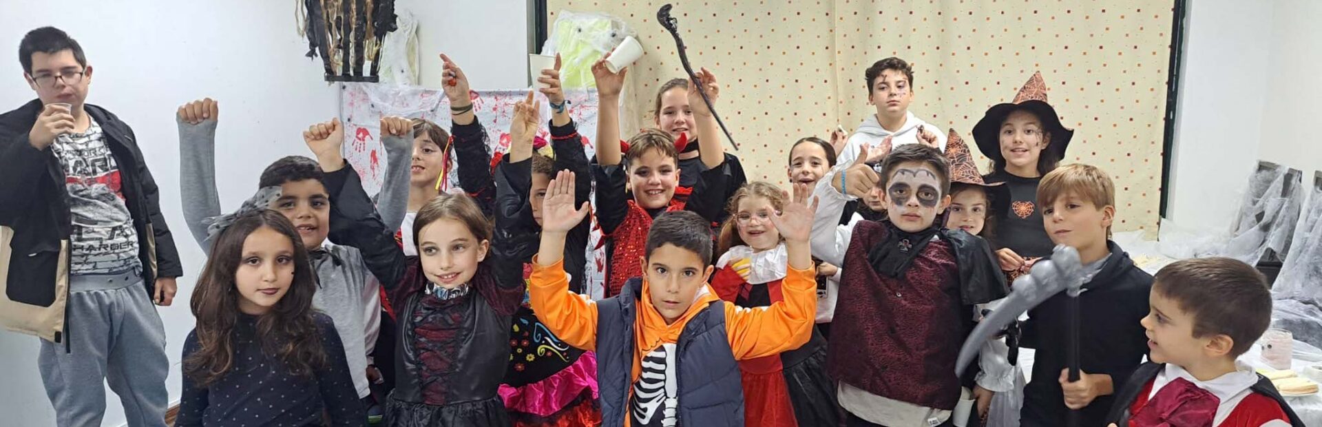 Grupo de crianças e jovens em sala interior. Estão praticamente todos mascarados com algo alusivo ao Dia das Bruxas.