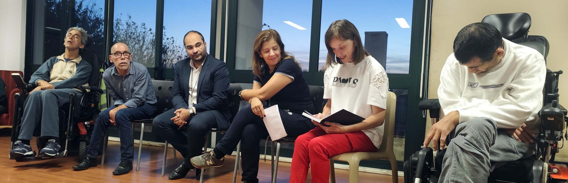 Grupo de seis pessoas sentadas em sala interior – sendo que duas usam cadeira de rodas. Uma das pessoas aparenta estar a falar enquanto lê algo que estará escrito num pequeno caderno.