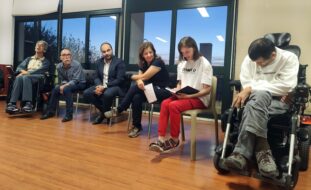 Grupo de seis pessoas sentadas em sala interior – sendo que duas usam cadeira de rodas. Uma das pessoas aparenta estar a falar enquanto lê algo que estará escrito num pequeno caderno.