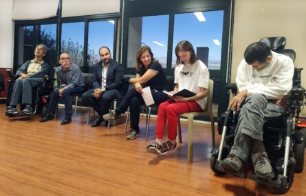 Grupo de seis pessoas sentadas em sala interior – sendo que duas usam cadeira de rodas. Uma das pessoas aparenta estar a falar enquanto lê algo que estará escrito num pequeno caderno.