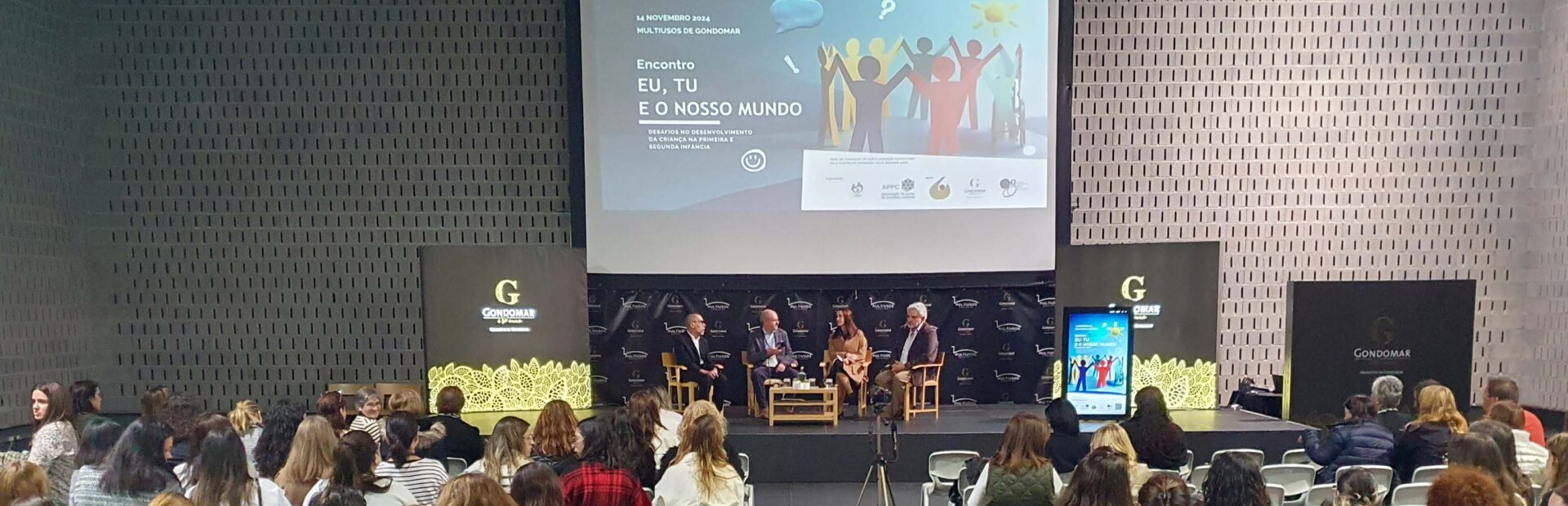Fotografia de espaço interior amplo. Mais de uma centena de cadeiras com pessoas sentadas (de costas). Ao fundo um palco com quatro pessoas sentadas. Projeção em tela na parede atrás das pessoas e cartazes laterais.