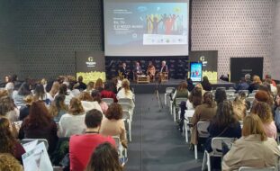 Fotografia de espaço interior amplo. Mais de uma centena de cadeiras com pessoas sentadas (de costas). Ao fundo um palco com quatro pessoas sentadas. Projeção em tela na parede atrás das pessoas e cartazes laterais.
