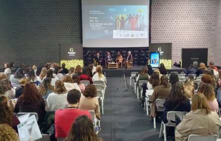 Fotografia de espaço interior amplo. Mais de uma centena de cadeiras com pessoas sentadas (de costas). Ao fundo um palco com quatro pessoas sentadas. Projeção em tela na parede atrás das pessoas e cartazes laterais.