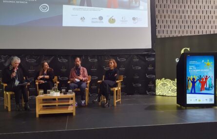 Fotografia de um palco com quatro pessoas sentadas junto de uma pequena mesa. Uma das pessoas segura um microfone e aparenta estar a falar. Projeção em tela na parede atrás das pessoas. Do lado direito da imagem um cartaz e uma projeção multimédia na parte frontal de um púlpito.