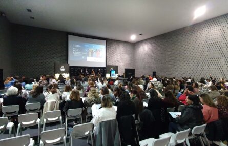 Fotografia de espaço interior amplo. Mais de uma centena de cadeiras com pessoas sentadas (de costas). Ao fundo um palco com quatro pessoas sentadas. Projeção em tela na parede atrás das pessoas e cartazes laterais.
