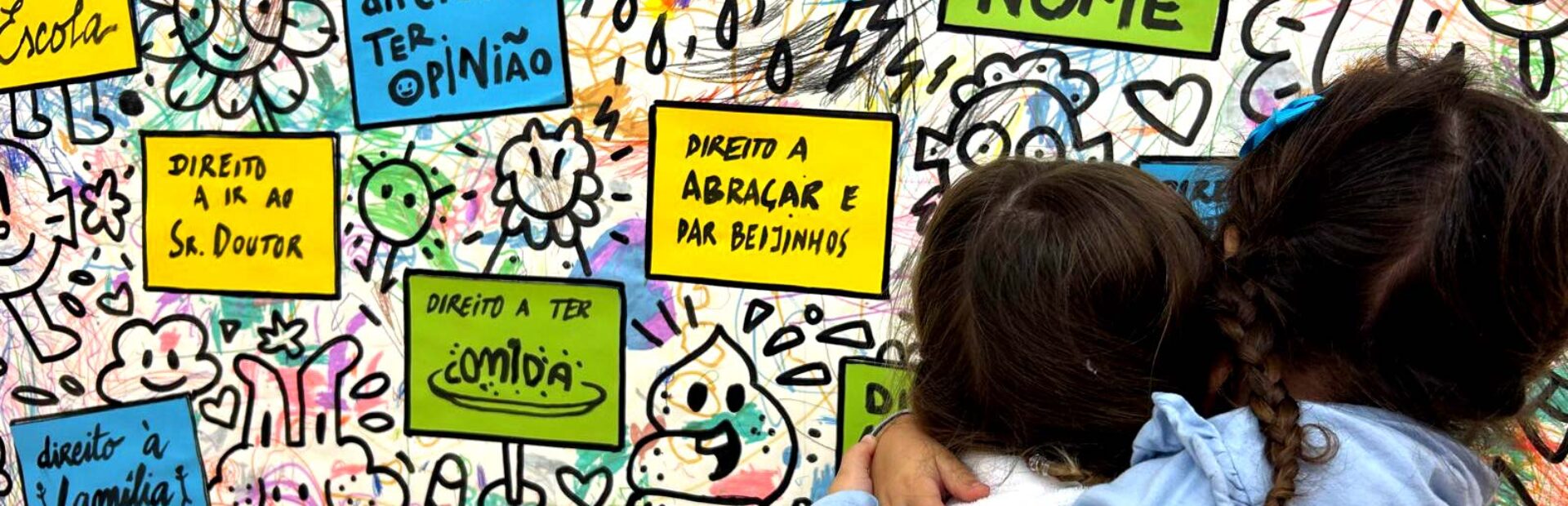 Duas crianças, de costas, abraçadas. Estão junto de um cartaz afixado numa parede – cartaz esse com imensas imagens, riscos coloridos e várias mensagens sobre os direitos das crianças.