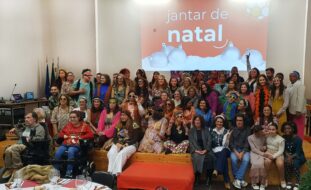 Fotografia de grupo com mais de meia centena de pessoas. Muitas das pessoas usam acessórios e roupas antigas (anos 70 do século passado). Atrás do grupo, em projeção multimédia na parede, uma imagem em tons de vermelho e branco com a expressão 