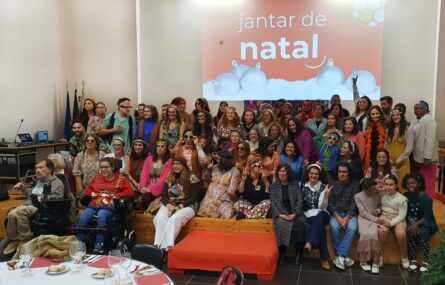 Fotografia de grupo com mais de meia centena de pessoas. Muitas das pessoas usam acessórios e roupas antigas (anos 70 do século passado). Atrás do grupo, em projeção multimédia na parede, uma imagem em tons de vermelho e branco com a expressão 