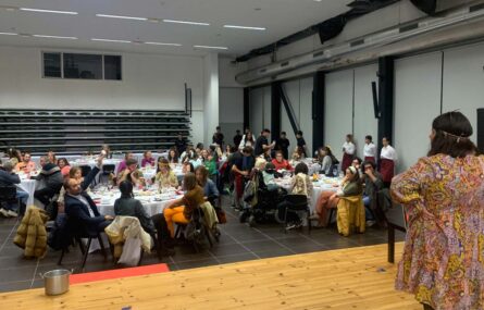 Imagem de sala espaçosa com várias mesas e inúmeras pessoas sentadas junto das mesmas. Em primeiro plano, no palco, uma pessoa de costas.