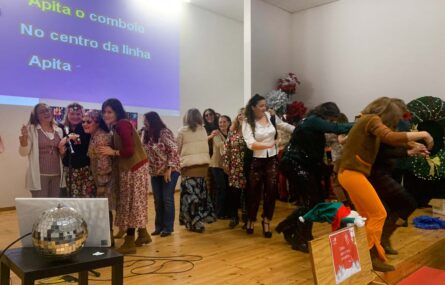 Perto de duas dezenas de pessoas em palco. Aparentam dançar e fazer uma fila. Quatro das pessoas estão junto de um microfone (supostamente a cantar). Na parede projeção com a frase 