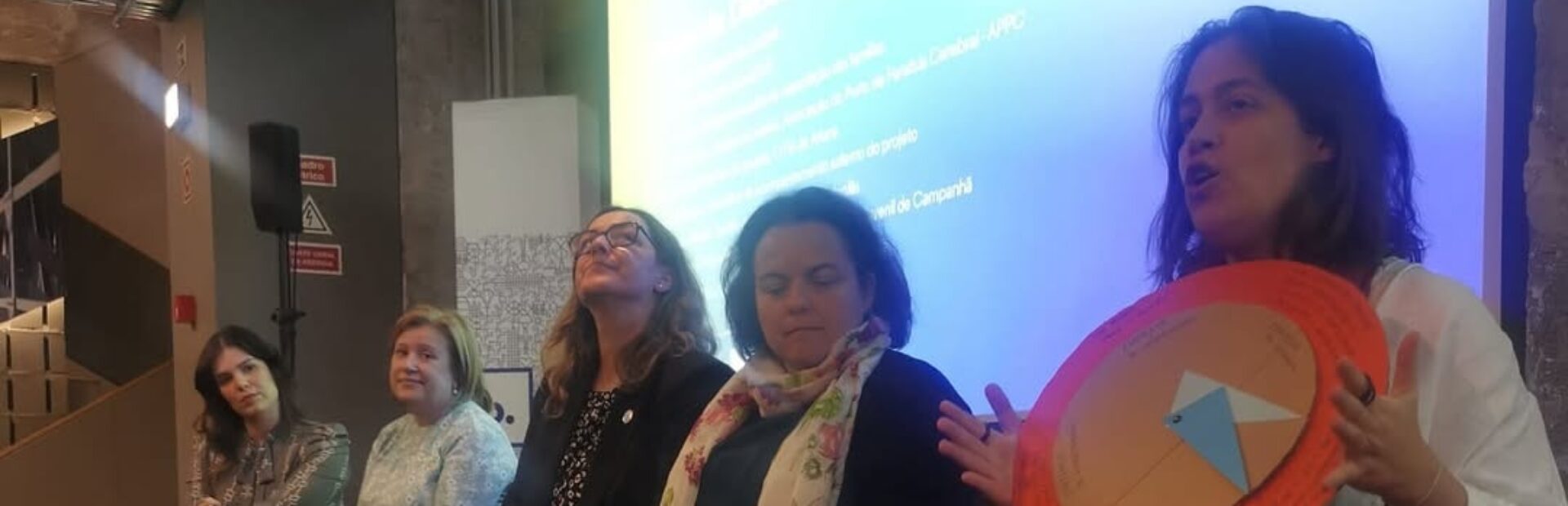 5 pessoas em palco com projeção atrás, uma das pessoas segura um objeto e dirige-se à plateia