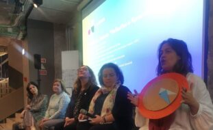 5 pessoas em palco com projeção atrás, uma das pessoas segura um objeto e dirige-se à plateia