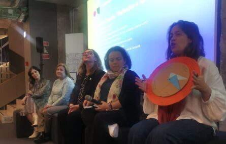 5 pessoas em palco com projeção atrás, uma das pessoas segura um objeto e dirige-se à plateia