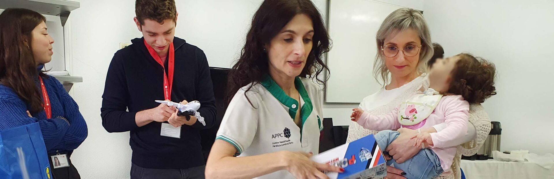 Imagem de quatro pessoas adultas e uma criança. Junto do grupo está uma mesa com vários brinquedos e caixas de brinquedos. O rosto da criança foi digitalmente desfocado.