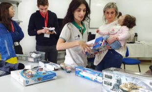 Imagem de quatro pessoas adultas e uma criança. Junto do grupo está uma mesa com vários brinquedos e caixas de brinquedos. O rosto da criança foi digitalmente desfocado.