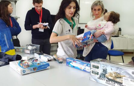 Imagem de quatro pessoas adultas e uma criança. Junto do grupo está uma mesa com vários brinquedos e caixas de brinquedos. O rosto da criança foi digitalmente desfocado.