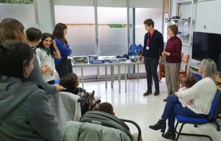 Imagem em sala interior de oito pessoas adultas e três crianças. Junto do grupo estão duas mesas com vários brinquedos e caixas de brinquedos.