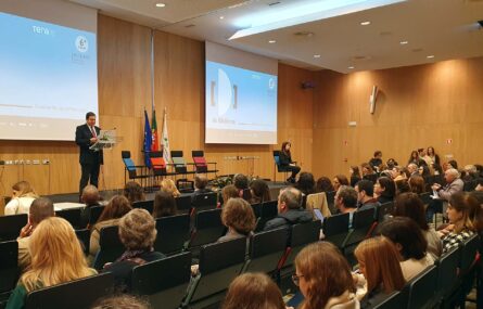 Imagem de auditório interior. Várias dezenas de pessoas sentadas em várias filas de cadeiras. Ao fundo, em palco, uma pessoa junto de um púlpito. Atrás dessa pessoa, na parede, duas projeções multimédia em telas.