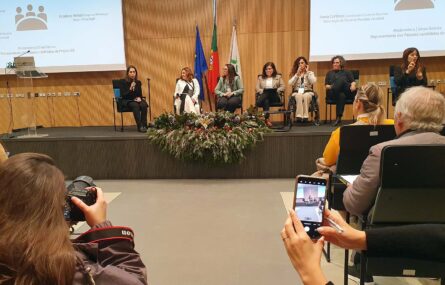 Em primeiro plano, de costas, duas pessoas fotografam (utilizando máquina fotográfica e telemóvel). Ao fundo, em palco, sete pessoas sentadas. Atrás delas, na parede, duas projeções multimédia em telas.