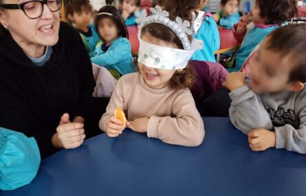 Uma criança, de olhos vendados, prepara-se para comer o que aparenta ser um gomo de laranja (ou tangerina). Outras crianças assistem, bem como uma pessoa adulta. Os rostos visíveis das crianças foram digitalmente desfocados.