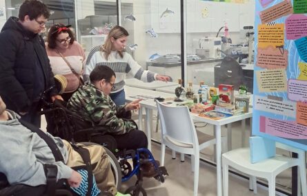 Cinco pessoas junto de uma mesa com diversos produtos. Ao lado está um quadro repleto de pequenos textos. Atrás de paredes em vidro vê-se o que aparenta ser um laboratório com diverso equipamento.