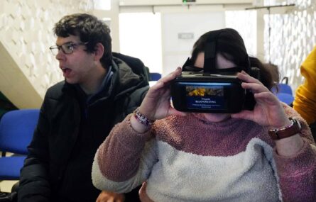 Duas pessoas sentadas. Uma das pessoas tem colocado na cabeça um equipamento que aparenta ser para visualização de um vídeo. No equipamento digital lê-se “Projeto BlueForesting”.
