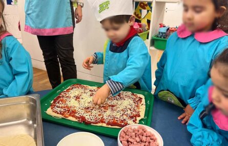 Quatro crianças junto de uma mesa. Uma das crianças espalha pequenos pedaços de queijo e de fiambre em cima de uma pizza ainda crua. Os rostos visíveis das crianças foram digitalmente desfocados.