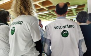 Plano aproximado de duas pessoas, de costas. Ambas usam t-shirts brancas com o logótipo “Bel” e a palavra voluntário. São ainda visíveis mais algumas pessoas na imagem.