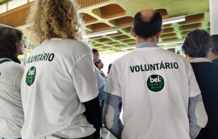 Plano aproximado de duas pessoas, de costas. Ambas usam t-shirts brancas com o logótipo “Bel” e a palavra voluntário. São ainda visíveis mais algumas pessoas na imagem.