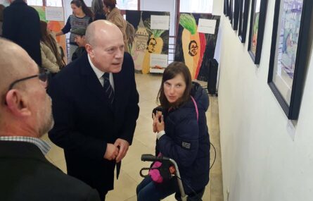 Várias pessoas em espaço interior. Em primeiro plano estão três pessoas que olham para um dos quadros expostos na parede. Ao fundo da imagem mais trabalhos artísticos em exposição.