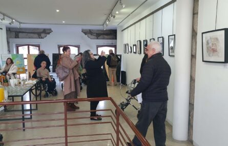 Espaço interior com vários trabalhos em exposição. Uma dezena de pessoas a circular pela sala. Do lado esquerdo da imagem uma mesa com vários copos e alguns produtos comestíveis indistintos.