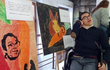 Uma pessoa junto de dois quadros coloridos que apresentam abordagens artísticas de um auto-retrato. Os quadros apresentam ainda curtos textos escritos.