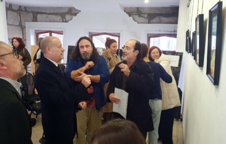 Várias pessoas em espaço interior. Um pequeno grupo de três pessoas aparenta conversar enquanto gesticulam. Na parede direita estão vários quadros expostos.
