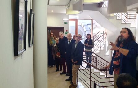 Várias pessoas em espaço interior. Em destaque surge uma pessoa que aparenta estar a fazer alguma explicação sobre um dos quadros em exposição. As restantes pessoas aparentam ouvir o que é dito. Na parede são parcialmente visíveis dois quadros.