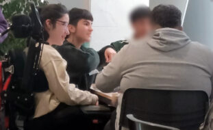Grupo de quatro jovens junto de uma mesa em sala interior. Um dos rostos foi digitalmente desfocado.