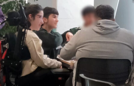 Grupo de quatro jovens junto de uma mesa em sala interior. Um dos rostos foi digitalmente desfocado.