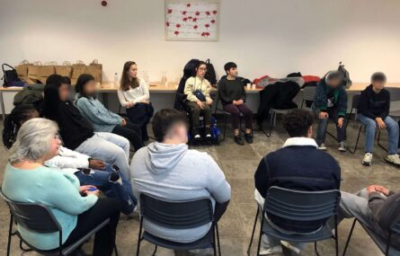 Grupo de 12 jovens e adultos sentados em círculo em espaço interior (sala ampla). Ao fundo, junto da parede, algumas mesas com material diverso. Na parede um quadro com vários cravos encarnados e brancos (aparentemente em papel). A maioria dos rostos (visíveis) foi digitalmente desfocada.