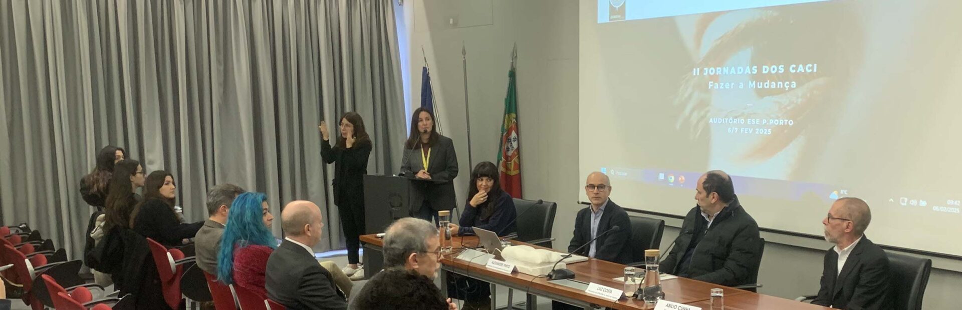 Imagem de auditório interior. Mais de uma dezena de pessoas sentadas na zona da plateia. Mesa com quatro pessoas sentadas junto da mesma. Uma pessoa fala junto de um púlpito enquanto outra aparenta fazer tradução para Língua Gestual Portuguesa. Projeção multimédia ao fundo da imagem.