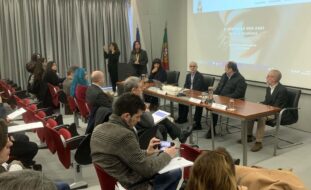 Imagem de auditório interior. Mais de uma dezena de pessoas sentadas na zona da plateia. Mesa com quatro pessoas sentadas junto da mesma. Uma pessoa fala junto de um púlpito enquanto outra aparenta fazer tradução para Língua Gestual Portuguesa. Projeção multimédia ao fundo da imagem.