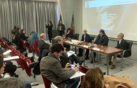 Imagem de auditório interior. Mais de uma dezena de pessoas sentadas na zona da plateia. Mesa com quatro pessoas sentadas junto da mesma. Uma pessoa fala junto de um púlpito enquanto outra aparenta fazer tradução para Língua Gestual Portuguesa. Projeção multimédia ao fundo da imagem.
