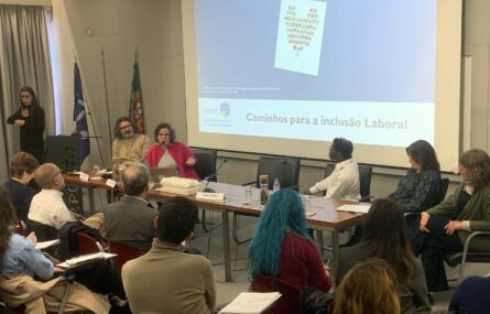 Imagem de auditório interior. Mais de uma dezena de pessoas sentadas na zona da plateia. Mesa com cinco pessoas sentadas junto da mesma. Uma pessoa junto de um púlpito aparenta fazer tradução para Língua Gestual Portuguesa. Projeção multimédia ao fundo da imagem.