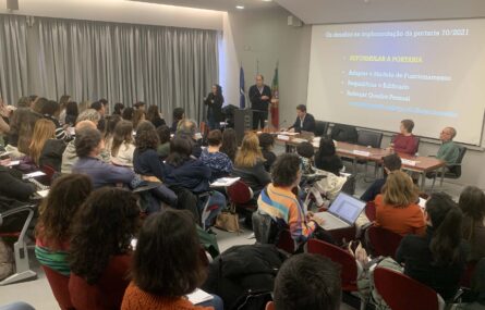 Imagem de auditório interior. Várias dezenas de pessoas sentadas na zona da plateia. Mesa com três pessoas sentadas junto da mesma. Uma pessoa junto de um púlpito aparenta estar a falar (enquanto outra, ao lado, parece estar a fazer tradução para Língua Gestual Portuguesa). Projeção multimédia ao fundo da imagem.