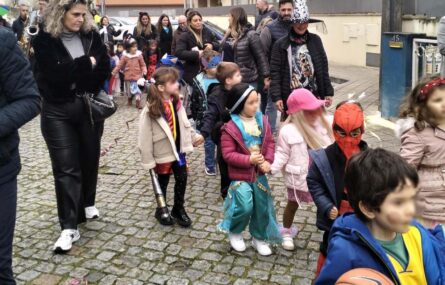 Grupo de crianças mascaradas em percurso por uma rua de paralelos. São acompanhadas por algumas pessoas adultas. Os rostos identificáveis das crianças foram digitalmente desfocados.