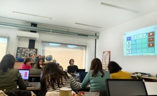Fotografia de sala de formação com diversas mesas, cadeiras e material informático. Na imagem surgem 11 pessoas e vê-se uma projeção multimédia na parede.