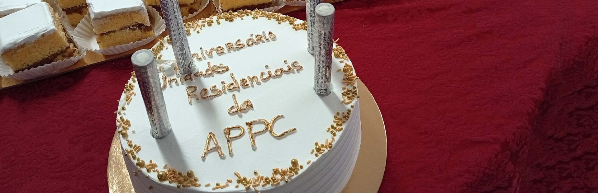 Fotografia de um bolo de aniversário, redondo, pousado numa mesa com uma toalha de tom roxo. Junto do pequeno bolo também várias pequenas fatias, em formato quadrado, de um bolo previamente cortado.